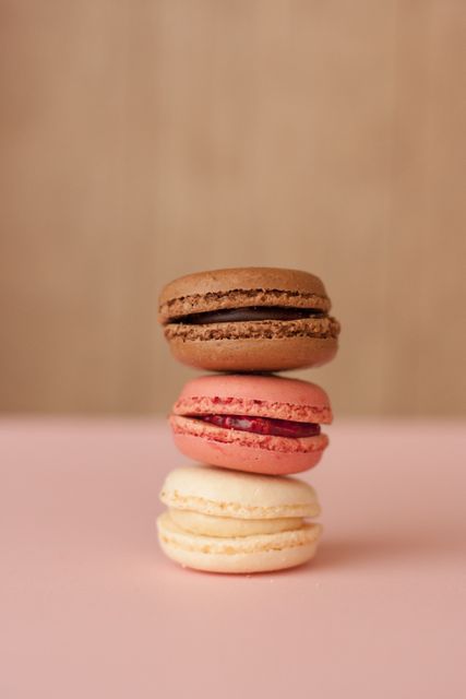 Three macarons in chocolate, raspberry, and vanilla flavors stacked on a pastel pink surface with a blurred, neutral background. This colorful and mouth-watering image is perfect for use in bakery advertisements, recipe books, food blogs, and cafe promotions highlighting desserts and confections.