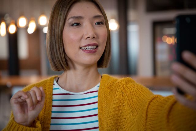 Biracial female using smartphone for a video call in a modern office. She is wearing a yellow sweater and striped shirt, smiling and engaging in conversation. Ideal for use in business communication, remote work, technology, and modern office environment themes.