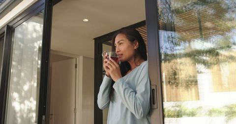 Mid-adult asian woman sipping tea by sliding glass door, enjoying morning sunlight and calm