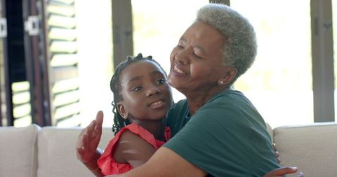 Senior african grandmother embracing granddaughter on sunlit sofa at home