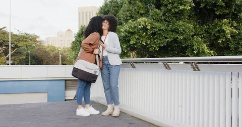 Two women exchanging cheek kisses on city bridge, casual friendship with skyline backdrop