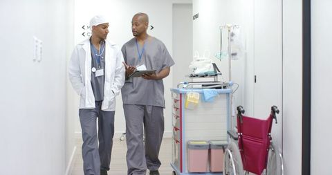African american clinicians walking hospital corridor discussing patient notes on tablet