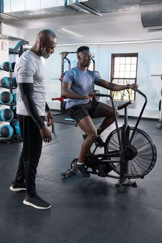 African american man powering air bike with personal trainer coaching cardio and strength