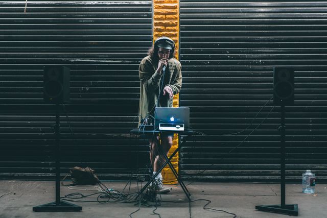 DJ performing live set on street with industrial backdrop, laptop and audio equipment. Ideal for projects on urban music culture, nightlife, street performances, and entertainment.
