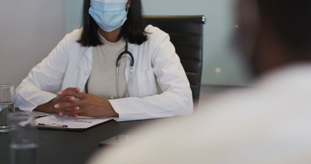 Female doctor wearing mask consulting with a patient in her office. A stethoscope is visible around her neck, indicating a medical setting. A clipboard with documents is placed on the table. Suitable for themes related to healthcare, medical consultations, safe practices during Covid-19, doctor's appointments, and medical assessments.