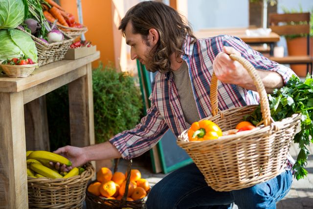 Man shopping for fresh fruits and vegetables in an organic market. He is holding a wicker basket filled with produce and selecting bananas from a display. This image is ideal for promoting healthy lifestyles, organic food markets, local produce, and sustainable living. It can be used in advertisements, blog posts, and articles related to healthy eating, organic farming, and local markets.