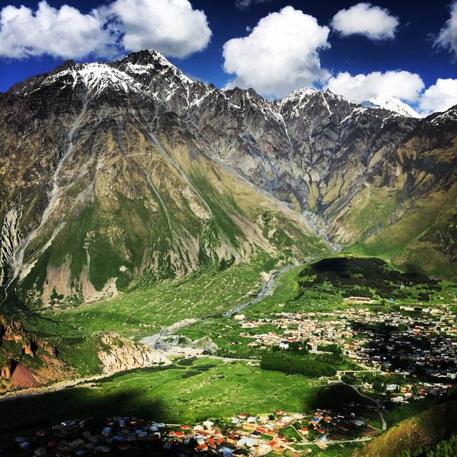 This serene mountain village is nestled at the base of towering snow-capped peaks under a partly cloudy sky. It features lush greenery and divided streams carving through the valleys, presenting a stunning contrast of natural beauty. Ideal for promoting ecotourism, adventure travel, nature retreats, and outdoor activities. Perfect for use in travel guides, postcards, and nature magazines.