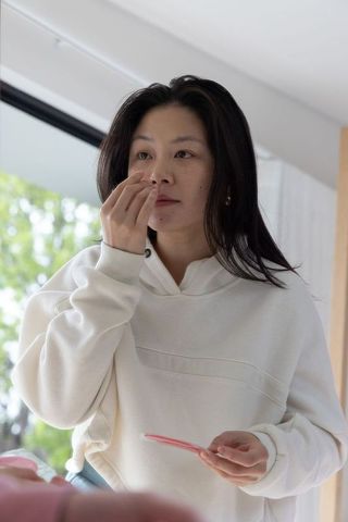 Asian woman touching nose holding pink compact mirror by window in white hoodie