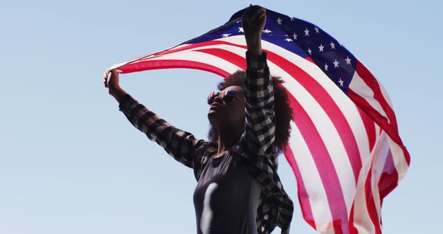 Woman outdoors holding American flag above head with both hands, waving gently in the breeze. She looks proud, joyful, and celebratory, representing values of freedom and independence. Great for concepts of national pride, patriotic holidays like Independence Day, Veterans Day, and Memorial Day. Suitable for advertisements, promotional materials, and educational uses.