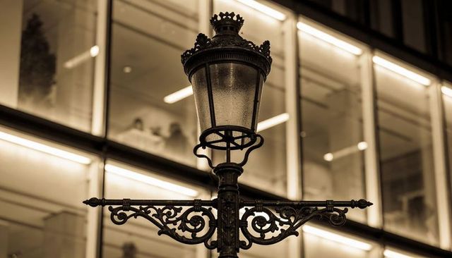Ornate cast-iron street lamp casting warm sepia glow against modern glass facade at night
