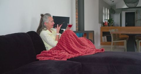 Senior woman relaxing on sofa with plush blanket and wine speaking into smartphone at home