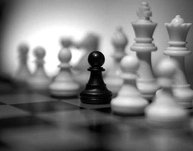 Black pawn standing prominently in contrast with blurred white pieces on a chessboard. Useful for illustrating strategy, competition, and the concept of standing out or being different.