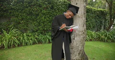 African american graduate wearing gown and mortarboard writing in ribboned notebook outdoors