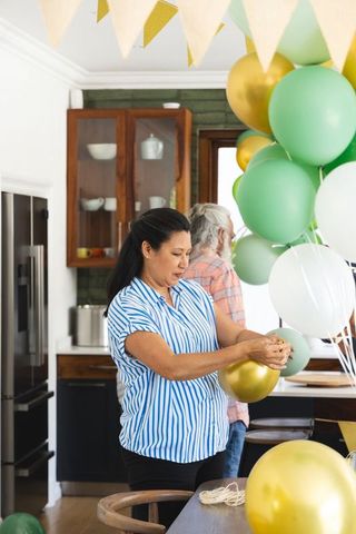 Mature indian man and diverse woman decorating kitchen with gold and green balloons