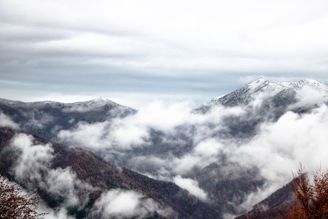 A serene view of misty mountains with snow-covered peaks enveloped in clouds under a cloudy sky. Suitable for travel magazines, nature blogs, wallpapers, or environmental campaigns to evoke a sense of adventure, tranquility, and the beauty of nature's winter landscapes.