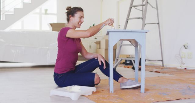 Woman painting a piece of furniture with a brush in newly renovated home. She is sitting on the floor in casual clothes, smiling and focusing on her task. Room is spacious and bright, with a ladder and boxes in the background indicating ongoing home projects. Ideal for content related to DIY enthusiasts, home renovation, creative activities, and personal projects.