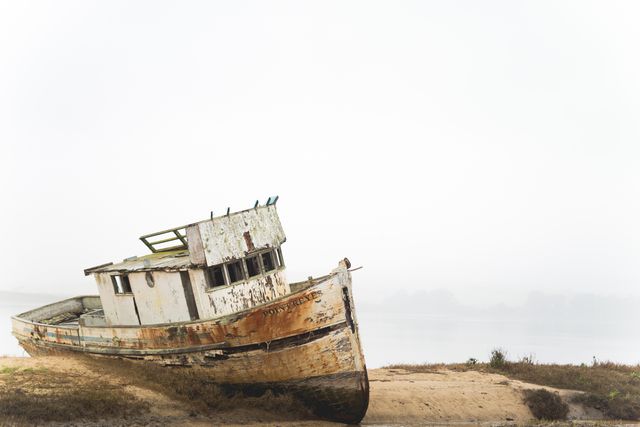 A derelict fishing boat is rusting away on a fog-shrouded coast, partially buried in the sand, creating an evocative scene of maritime abandonment. Ideal for themes of nostalgia, decay, and maritime history, this image can be used for coastal travel blogs, nautical-themed articles, and artistic projects focusing on weathered and rustic aesthetics.