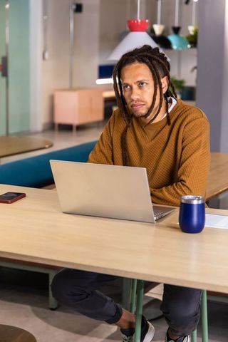 African American man working on laptop at coworking table, listening with earbuds, focused