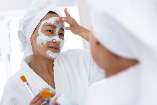Happy asian woman looking in bathroom mirror, putting on beauty face mask, copy space. Self care, health, relaxation, inclusivity and lifestyle.