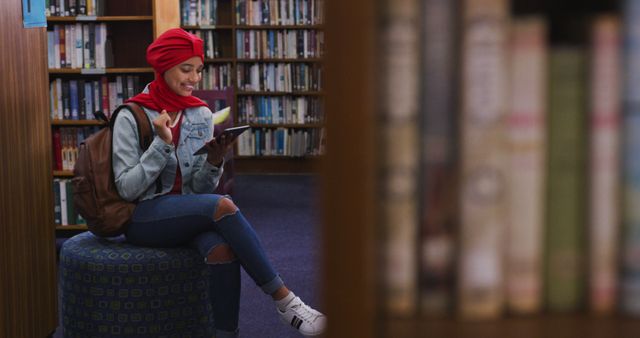 A young woman wearing a headscarf and casual clothing, including a denim jacket and jeans, sits on a pouf in a library while reading an e-book and smiling. She has a backpack and appears engaged in her reading. Ideal for illustrating themes such as education, technology in learning, leisure reading, or youth lifestyle in academic environments.
