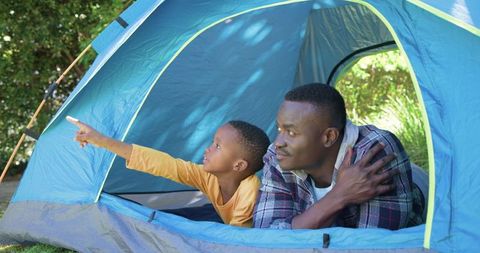 African american father and son camping in blue tent pointing toward sunny backyard
