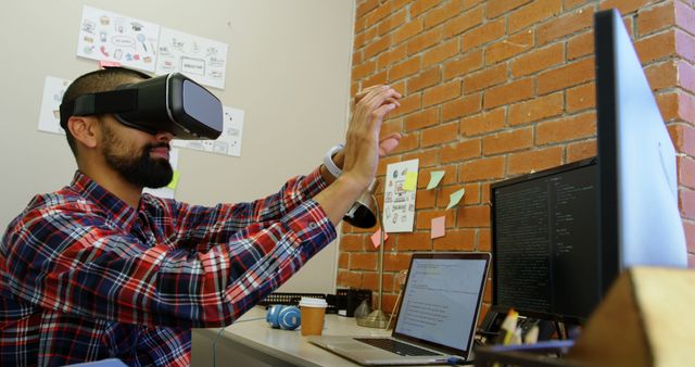 Software developer using virtual reality headset working on project in office environment. Coding setup with multiple screens and a laptop, surrounded by sticky notes and tech accessories. Ideal for illustrating tech workplaces, innovative work methods, and the integration of virtual technology in professional settings.