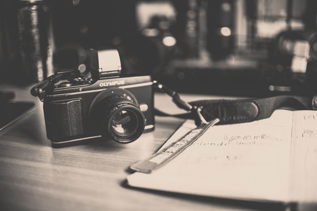 This image shows a vintage Olympus camera placed on a wooden desk next to sketchbooks containing drawings. The black and white tone gives it a retro feel, making it ideal for use in topics related to creativity, artistic inspiration, photography, and retro aesthetics. Useful for publications on photography, artistic blogs, tutorials on drawing or sketching, and vintage technology.