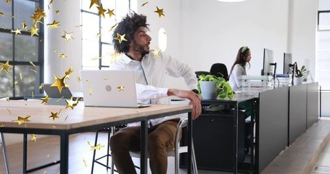 Middle eastern man celebrating with gold confetti in modern coworking office at wooden desk