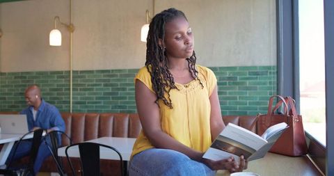African woman reading paperback in sunlit cafe window wearing yellow blouse and jeans