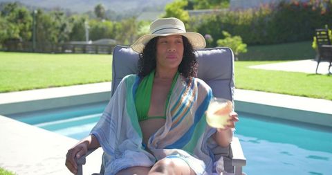 African american woman relaxing by pool in straw hat holding refreshing drink on patio