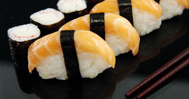 A selection of sushi, including nigiri topped with slices of fresh salmon and maki rolls, is presented elegantly on a dark, reflective surface. Chopsticks rest beside the sushi, inviting the viewer to indulge in the traditional Japanese cuisine.