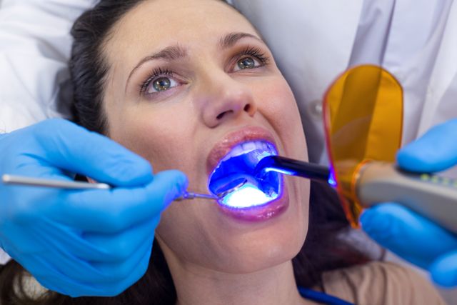 Dentists examining female patient using dental curing light in a clinic. Ideal for use in articles or advertisements related to dental care, oral health, dental clinics, and professional dental services. Can also be used in educational materials about dental procedures and hygiene.