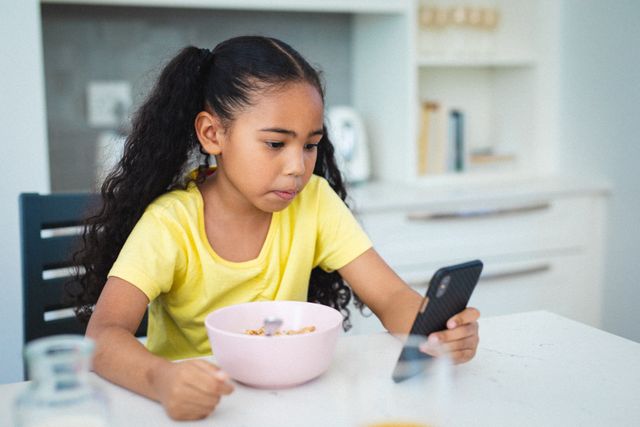 Hispanic girl sitting at dining table in kitchen, using smartphone while eating breakfast. Perfect for illustrating childhood, technology use, morning routines, and modern family life. Useful for articles on screen time, healthy eating habits, and balancing technology with daily activities.