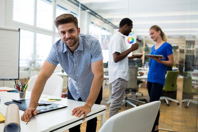Male graphic designer smiling confidently while leaning on desk in modern office. Coworkers in background interacting and collaborating on project. Ideal for use in business, teamwork, and creative industry contexts, showcasing a positive and productive work environment.