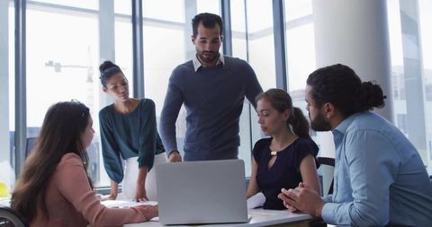 Business team collaborating around laptop in modern glass office, leader guiding discussion