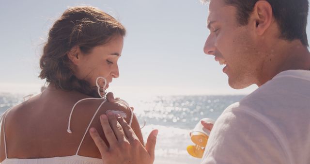 A young Caucasian couple enjoys a sunny beach setting, with the woman wearing a white dress and the man in a casual white shirt. Their relaxed and intimate posture suggests a romantic connection as they savor a moment by the sea.