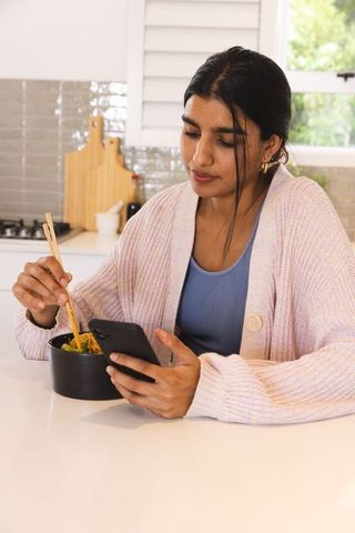 Young woman using phone while enjoying noodle meal in kitchen