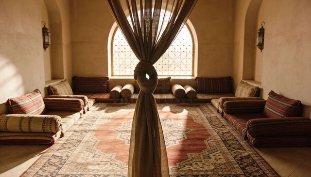 Knotted curtain revealing sunlit majlis with arched lattice window and patterned rug