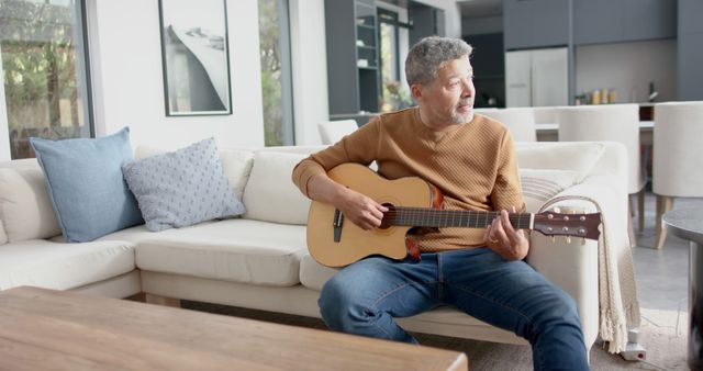 Middle-aged man casually playing an acoustic guitar while sitting on a sofa in a modern and stylish living room. Ideal for concepts related to home entertainment, music practice, relaxed lifestyle, leisure time, and modern home interiors. Use in articles, blogs, or advertisements focused on hobbies, relaxation, music lessons, or home decor.