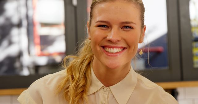 Young woman smiling in a brightly lit cafe, giving warm, friendly vibe. Perfect for use in advertisements focusing on social interactions, hospitality industry promotion, or online content emphasizing positivity and approachability.