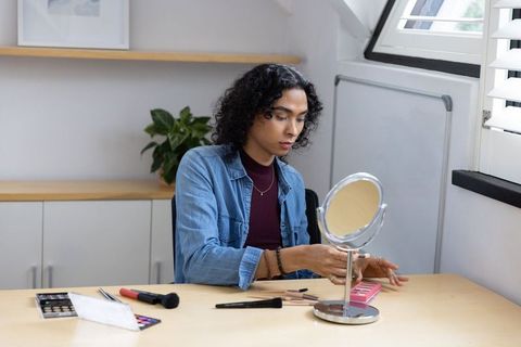 Non-binary person applying eyeshadow at desk by window with mirror and pink palette