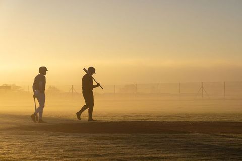 Teammates walking across misty baseball field at golden hour carrying bats and helmets