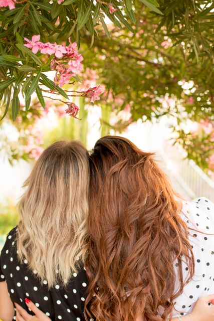 Two women with long, flowing hair are shown from behind, with one woman having blonde hair and the other having red hair. They are embracing under a blossoming tree, surrounded by greenery and pink flowers. This image symbolizes friendship, togetherness, and the beauty of nature, perfect for use in social media, blog posts, and articles about friendship, outdoor activities, or springtime.