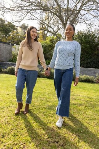 Diverse female couple walking hand-in-hand across sunlit backyard in casual sweaters