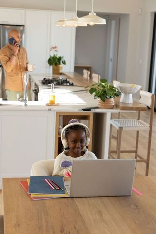African american father talking on phone while daughter studying on laptop kitchen table