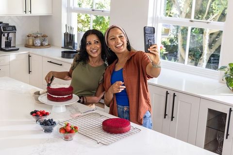 Friends frosting red velvet cake and taking selfie in bright modern kitchen