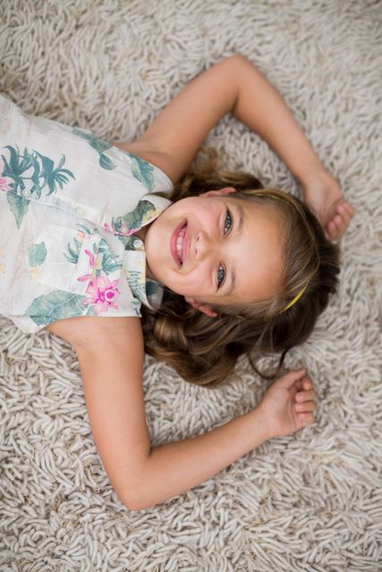 Young girl lying on a soft rug in a living room, smiling and looking at the camera. Ideal for use in advertisements, family lifestyle blogs, children's product promotions, and home decor magazines.