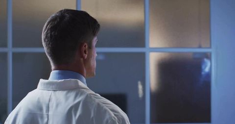 Researcher standing at frosted glass peering into sterile laboratory corridor with soft blue lightin