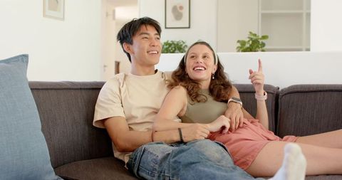 Young couple relaxing on sofa, smiling and pointing upward, modern home intimacy