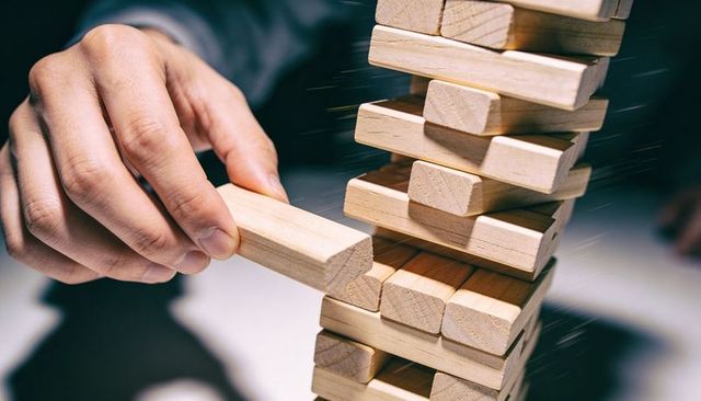Hand pulling wooden block from wobbly tower representing risk strategy and tension